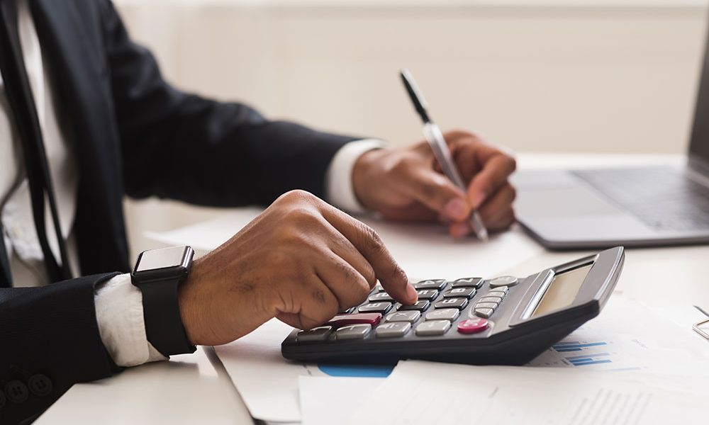 Person calculating finances with a calculator and laptop in an office setting, focusing on business documents.