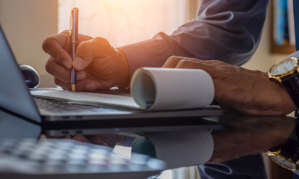Person writing notes with pen beside laptop on a desk, sunlight filtering through window.