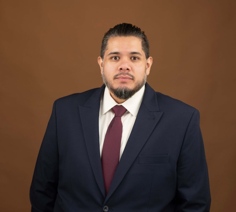 Juan Quintanilla in a suit and tie against a brown backdrop, looking into the camera.
