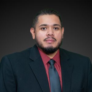 Professional man in a suit with a red shirt and gray tie, posing against a dark background.