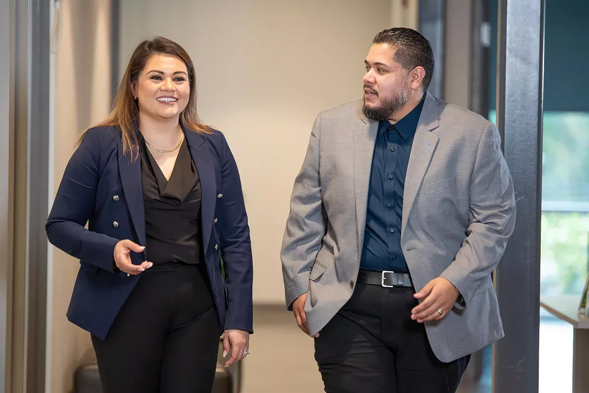Two professionals walking and talking in a modern office hallway, wearing business attire.