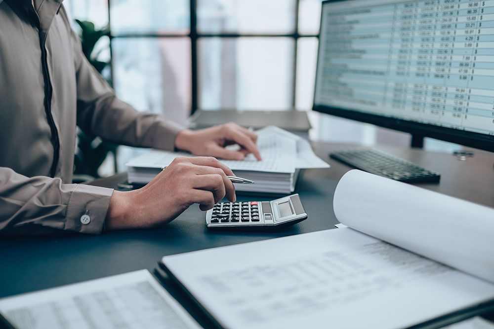 Person working on accounting with calculator and paperwork at desk, financial spreadsheets on computer screen.