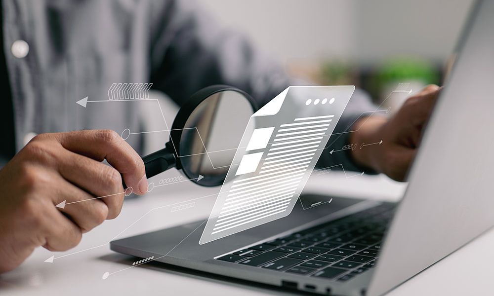 Person using magnifying glass to analyze digital documents on a laptop, symbolizing online data analysis and research.