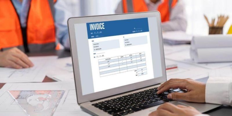 Laptop screen shows an invoice while two workers in orange safety vests work at a desk in the background.