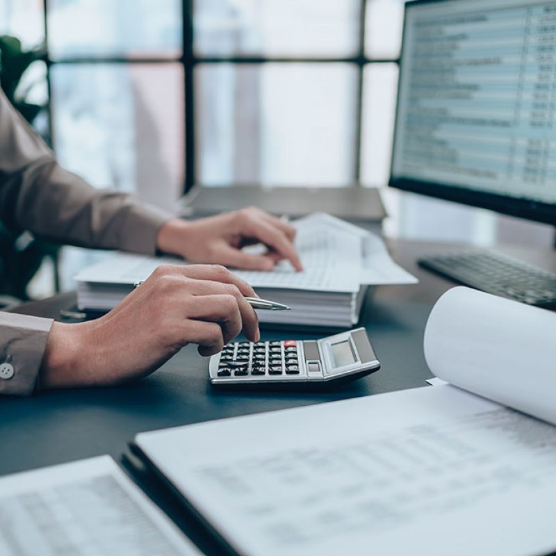 Person working on accounting with calculator and paperwork at desk, financial spreadsheets on computer screen.