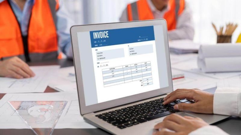 Laptop screen shows an invoice while two workers in orange safety vests work at a desk in the background.