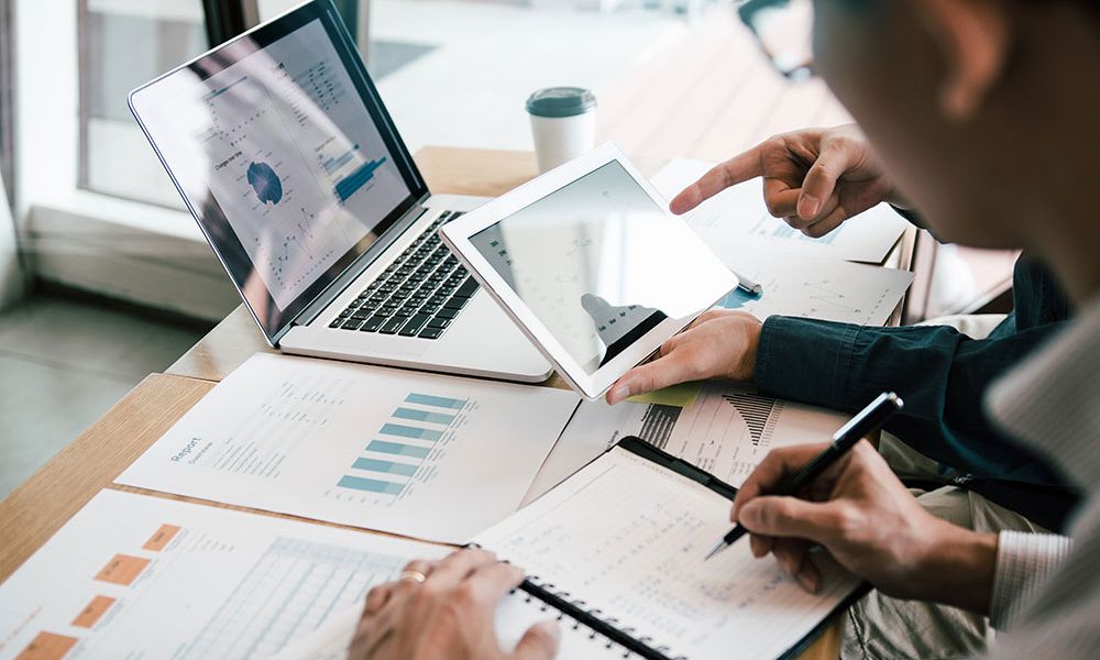 Team analyzing financial charts on laptop and tablet at office desk with notebooks and papers.