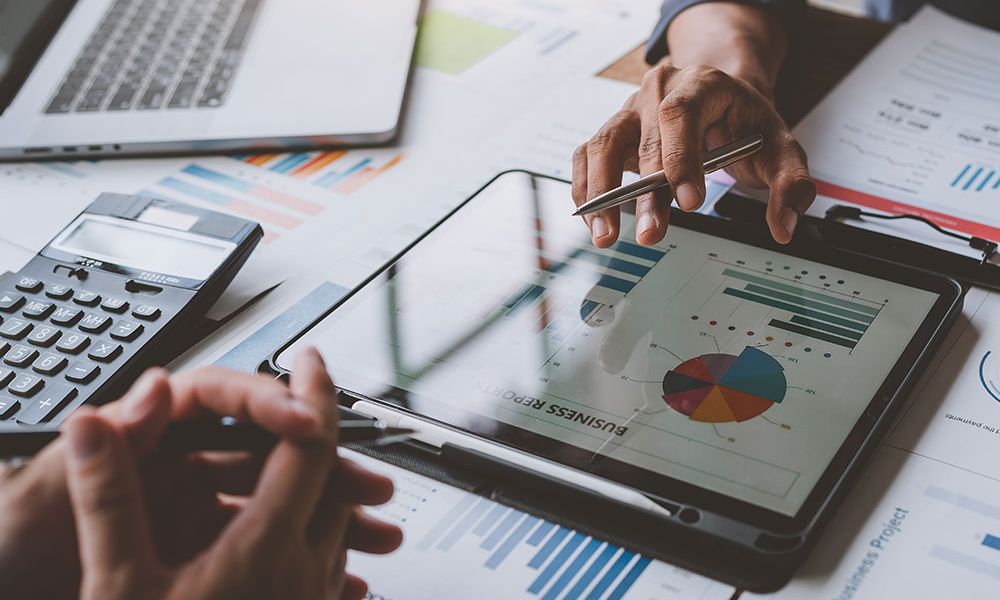 Business meeting analyzing charts on tablet and papers; laptop and calculator on table.