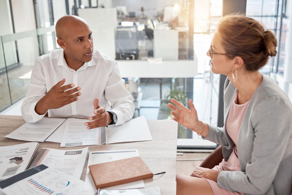 Two professionals discuss business strategy at a table with documents in a modern office setting.