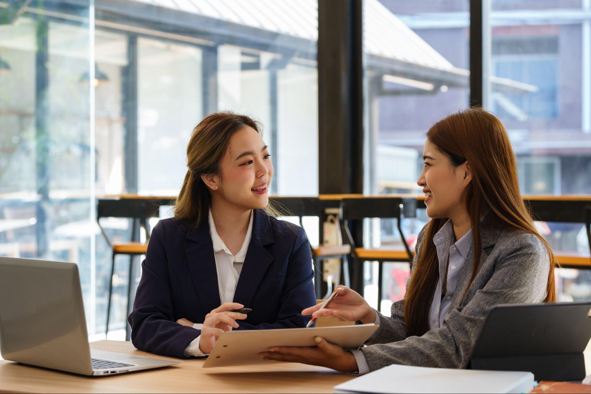 Two women in business attire discuss work at a table with laptops and documents in a bright office setting.