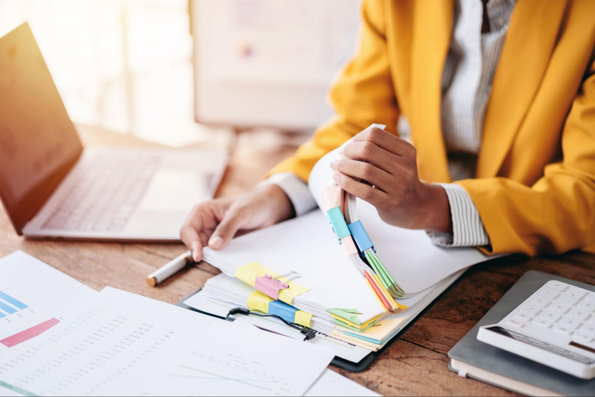 tax-planning – King of Kings Person in yellow jacket organizing documents at desk with laptop and calculator.