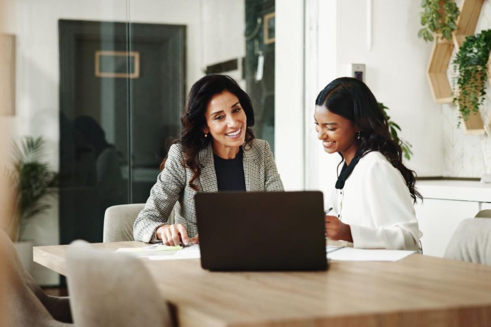 Two businesswomen collaborating at a laptop in a modern office space, smiling and discussing documents.