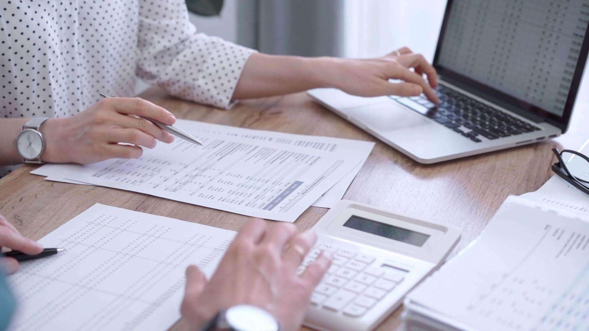 two people sitting at a table with papers and a laptop reviewing numbers